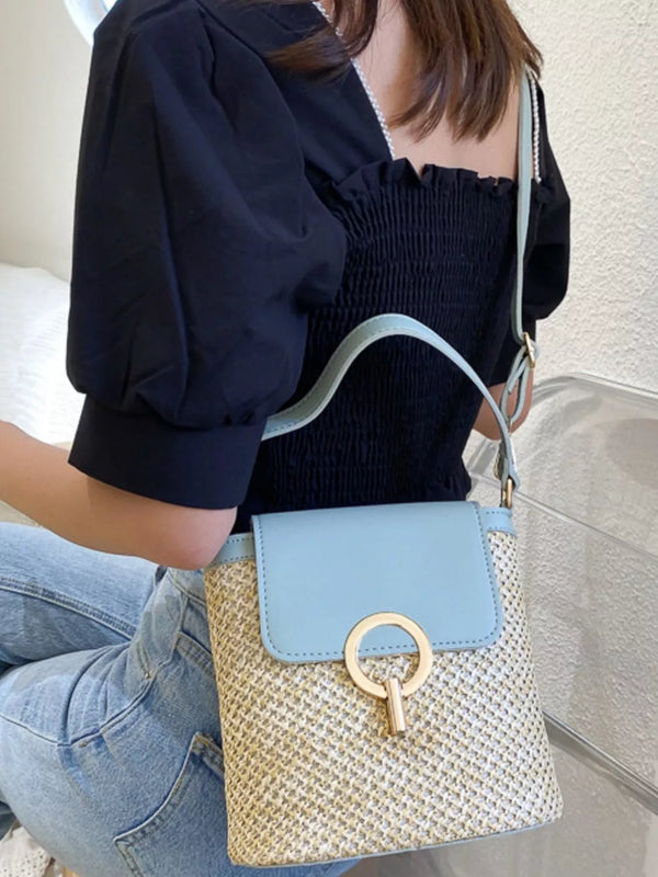 medium woven straw crossbody bag with blue flap and gold ring detail worn by woman in black top and jeans