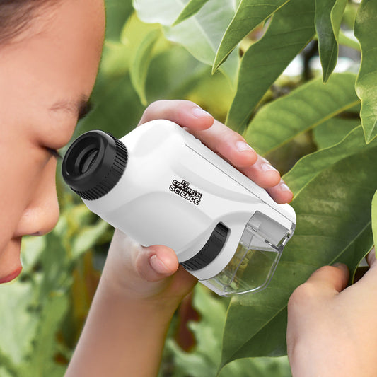 Child using a white handheld optical microscope to examine a leaf in nature.