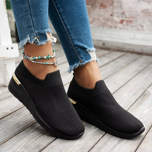Black mesh round toe wedge athletic shoes with mid heels and gold accent on sole worn with ripped blue jeans and ankle bracelets